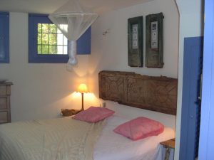 Dormitorio con cabecero de madera tallada, cojines rosas y contraventanas azules